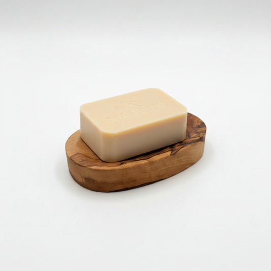 A beige rectangular bar of Nablus soap resting on a small oval wooden soap dish, set on a light gray surface.