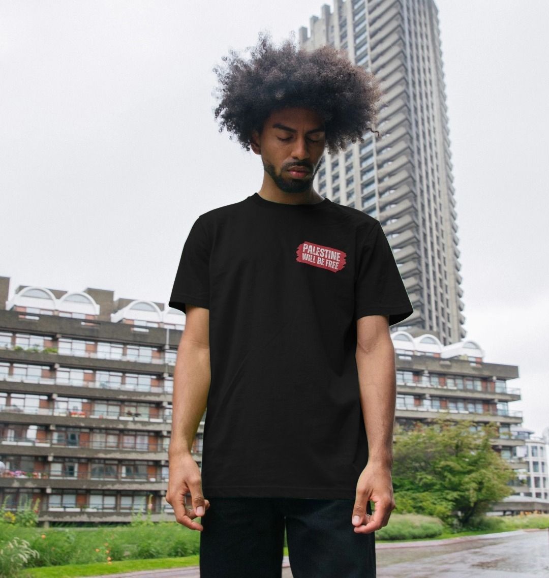 A man with an afro hairstyle wearing a black t-shirt with a red Palestine Will be Free graphic on the left chest, standing outdoors in front of modern apartment buildings under an overcast sky. 
