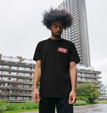 A man with an afro hairstyle wearing a black t-shirt with a red Palestine Will be Free graphic on the left chest, standing outdoors in front of modern apartment buildings under an overcast sky. 